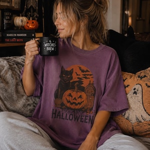 May include: A purple short-sleeve t-shirt featuring a graphic of a black cat, a jack-o'-lantern, a tombstone, and bats against an orange moon with the text "LONG LIVE HALLOWEEN" and a black mug with the text "WITCHES BREW".