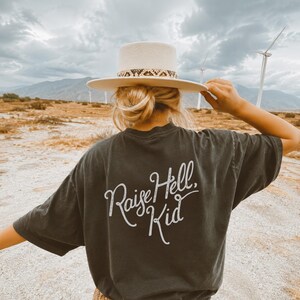 May include: A charcoal gray short-sleeve t-shirt featuring the white cursive text Raise Hell, Kid printed on the back. The shirt is paired with a cream-colored wide-brim hat that has a snakeskin-patterned band around the base of the crown.