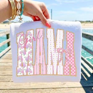 Puede incluir: Sudadera blanca con el texto "Girl Mama" en rosa, estampados florales y cuadros vichy. Las letras están delineadas con purpurina dorada. La sudadera es sostenida por una persona con pulseras doradas y una pulsera turquesa con forma de estrella de mar.