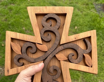 May include: A wooden cross with a carved design of leaves and swirls. The cross is made of two different types of wood, one light brown and one dark brown.