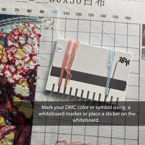 May include: A cross-stitch project with a white and black colour-coded tool. The tool has pink and blue threads attached. The text on the tool reads "3841". The text at the bottom says "Mark your DMC colour or symbol using a whiteboard marker or place a sticker on the whiteboard."