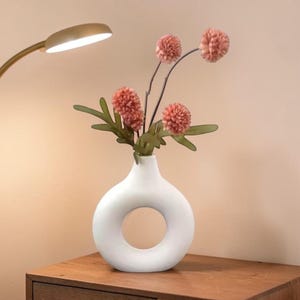 May include: A white ceramic vase with a circular opening holds pink flowers with green leaves. The vase sits on a wooden nightstand next to a gold-colored lamp.