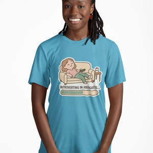 May include: A light blue t-shirt featuring a cartoon illustration of a person relaxing on a couch with a remote control. The text "INTROVERTING IN PROGRESS..." is displayed below the image, with a progress bar.