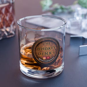 May include: A clear, lowball whiskey glass filled with amber liquid. The glass features a circular wooden-style label with the text "THE WHISKEY D.I.N.K'S" and social media icons. The glass sits on a dark surface.