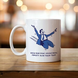 Könnte beinhalten: Weiße Keramik-Tasse mit einer blauen Illustration einer feiernden Person im Anzug. Der Text auf der Tasse lautet: "WHEN YOUR MINI LEAGUE RIVAL FORGETS TO DO THEIR TEAM."