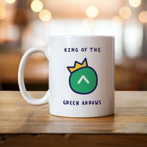 Könnte beinhalten: Weiße Keramiktasse mit dem Text "KING OF THE GREEN ARROWS". Die Tasse zeigt einen grünen Kreis mit einem weißen Pfeil und einem goldenen Kronen-Design. Die Tasse steht auf einer Holzoberfläche.