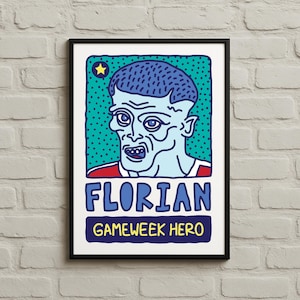 Könnte beinhalten: Ein gerahmter Kunstdruck mit einer Comic-Illustration einer Person mit blasser Haut, blauen Haaren und einem rot-weißem Hemd. Der Druck zeigt den Namen "FLORIAN" und den Text "GAMEWEEK HERO" in fetter Schrift.