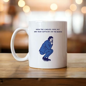 Könnte beinhalten: Weiße Keramik-Tasse mit einer blauen Illustration einer Person im Anzug, die hockt und ihr Gesicht bedeckt. Text über der Illustration: "WHEN THE LINEUPS COME OUT AND YOUR CAPTAIN'S ON THE BENCH."