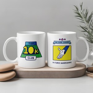 Könnte beinhalten: Zwei weiße Keramikbecher mit humorvollen Designs. Ein Becher zeigt eine grün-blaue Grafik mit dem Text "TOP 10K CLUB". Der andere Becher hat ein gelbes Quadrat mit einer Handschlag-Grafik und dem Text "YOU HAVE EARNED A FIRM HANDSHAKE."