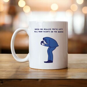 Könnte beinhalten: Weiße Keramik-Tasse mit Henkel. Auf der Tasse steht der Text "WHEN YOU REALISE YOU'VE LEFT ALL YOUR POINTS ON THE BENCH" und eine blaue Illustration einer Person, die nach unten schaut. Die Tasse steht auf einer Holzoberfläche.