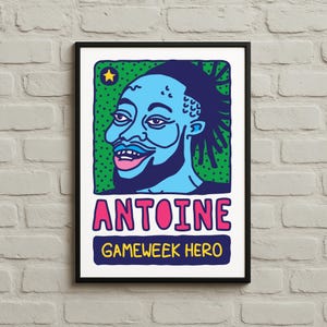 Könnte beinhalten: Ein gerahmter Kunstdruck mit einer stilisierten Illustration einer Person mit blauer Haut und Bart. Das Kunstwerk zeigt den Namen "ANTOINE" in Rosa über dem Text "GAMEWEEK HERO" in Gelb, vor einem grünen, gepunkteten Hintergrund.