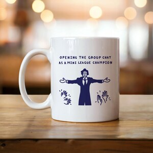 Könnte beinhalten: Weiße Keramiktasse mit einer blauen Grafik einer Person mit ausgebreiteten Armen und dem Text "OPENING THE GROUP CHAT AS A MINI LEAGUE CHAMPION". Die Tasse hat einen gebogenen Henkel und steht auf einer Holzoberfläche.