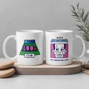 Könnte beinhalten: Zwei weiße Keramikbecher mit humorvollen Designs. Ein Becher zeigt eine grün-blaue Grafik mit "TOP 100K CLUB". Der andere Becher hat ein rosa Quadrat mit einem Gesicht und dem Text "YOU HAVE EARNED" und "AN APPROVING LOOK".