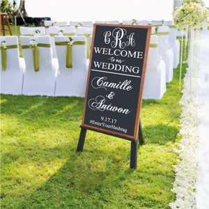 May include: A vertical rectangular chalkboard sign with a wooden frame on a black easel. The sign features white text reading CRA, WELCOME to our WEDDING, Camille & Antwon, 9.17.17, and #EnterYourHashtag in a mix of serif and script fonts.