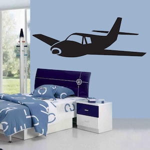 Könnte beinhalten: Eine schwarze Silhouette eines einmotorigen Flugzeugs vor einer hellblauen Wand. Das Flugzeug hat einen Propeller, Flügel und ein Heck. Das Bild ist Teil einer Schlafzimmerdekoration, mit Bett, Nachttisch und Fenster sichtbar.
