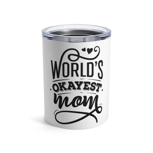 May include: A white cylindrical tumbler with a clear plastic lid featuring black text that reads World's Okayest Mom, accented with decorative swirls and two small hearts above the lettering. The design is centered on the front of the cup.