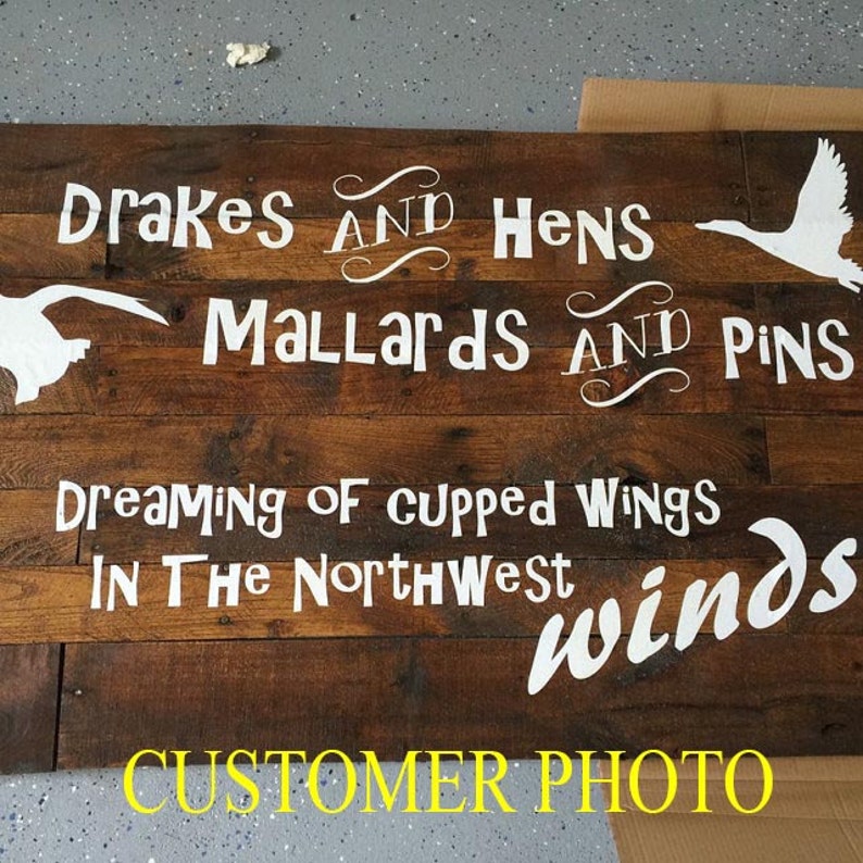 Op de afbeelding: Een rechthoekig houten bord gemaakt van donkerbruine horizontale planken met witte tekst: Drakes and Hens, Mallards and Pins, Dreaming of cupped wings in the Northwest winds, inclusief witte silhouetten van vliegende eenden.