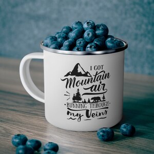 May include: White enamel mug filled with fresh blueberries. The mug has a silver rim and a black graphic that reads "I Got Mountain Air Running Through My Veins" with a mountain and bear illustration.
