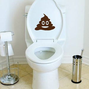 May include: A white toilet with a brown poop emoji decal on the lid. The toilet is in a bathroom with a white tiled floor and a chrome toilet paper holder.