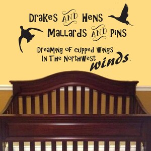 Op de afbeelding: Een zwarte muursticker met silhouetten van twee vliegende eenden en de tekst: Drakes and Hens, Mallards and Pins, Dreaming of cupped wings, In The Northwest winds. De sticker is aangebracht op een gele muur boven een donkerhouten ledikant.