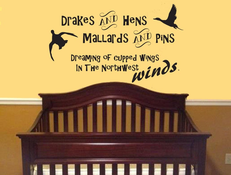 Op de afbeelding: Een zwarte muursticker met silhouetten van twee vliegende eenden en de tekst: Drakes and Hens, Mallards and Pins, Dreaming of cupped wings, In The Northwest winds. De sticker is aangebracht op een gele muur boven een donkerhouten ledikant.