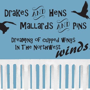 Op de afbeelding: Een muursticker met zwarte silhouetten van twee vliegende eenden en de tekst: Drakes AND Hens Mallards AND Pins Dreaming of cupped wings IN The Northwest winds. De sticker bevindt zich op een lichtblauwe muur boven een wit ledikant.