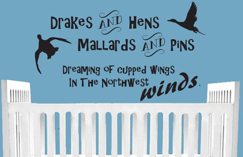 Op de afbeelding: Een muursticker met zwarte silhouetten van twee vliegende eenden en de tekst: Drakes AND Hens Mallards AND Pins Dreaming of cupped wings IN The Northwest winds. De sticker bevindt zich op een lichtblauwe muur boven een wit ledikant.