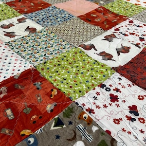 May include: A patchwork quilt with a variety of patterned squares. The quilt features squares with dog illustrations, cars, floral designs, and paw prints. Colors include red, green, white, and brown.