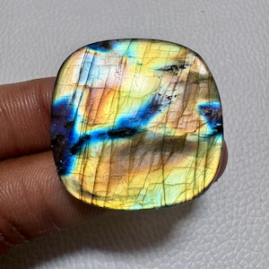 Puede incluir: Una piedra preciosa de labradorita de forma cuadrada con esquinas redondeadas. La piedra muestra un vibrante juego de colores, con tonos de azul, amarillo y naranja. La superficie tiene grietas visibles y un acabado pulido.