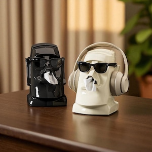 Puede incluir: Dos originales cajas de pañuelos con forma de estatuas Moai. Una es negra y la otra blanquecina. Ambas estatuas llevan gafas de sol y tienen pañuelos saliendo de la boca. La estatua blanquecina tiene auriculares.