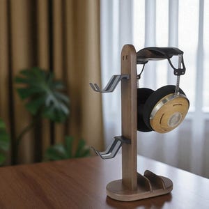 May include: A wooden headphone stand with two gray metal hooks and a curved top. A pair of black and wood-toned headphones hangs from the stand. The stand is on a wooden table.