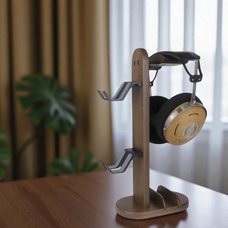 May include: A wooden headphone stand with two gray metal hooks and a curved top. A pair of black and wood-toned headphones hangs from the stand. The stand is on a wooden table.