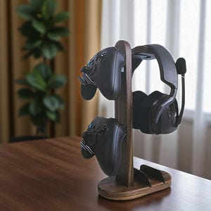 May include: A wooden gaming headset and controller stand. The stand is made of dark brown wood and holds two black controllers and a black headset with a microphone. The stand is on a wooden table.