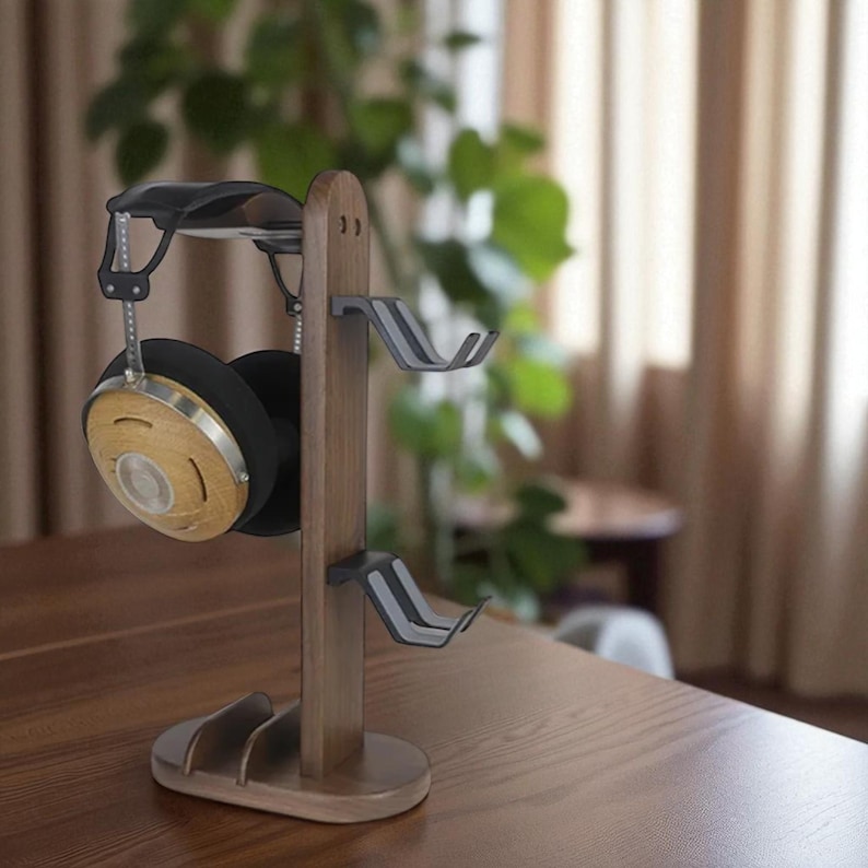 May include: A wooden headphone stand with a dark finish. The stand features a top rest for the headband and two side hooks for the earcups. A pair of headphones with wooden earcups and black padding are displayed on the stand. The stand is on a wooden table.