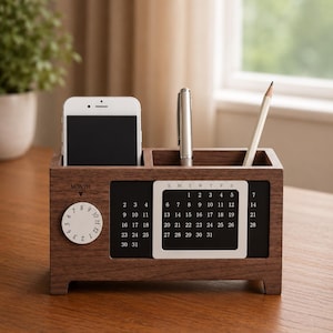 Op de afbeelding: Een rechthoekige bureau-organizer van donker hout met een ingebouwde eeuwigdurende kalender, een ronde maandwijzer en een dagrooster. De unit heeft twee vakken met een smartphone, een zilveren pen en een houten potlood.