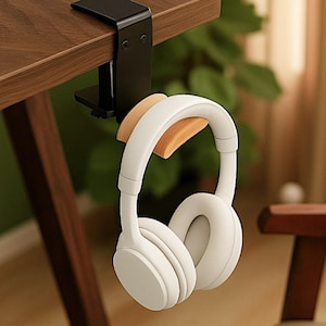 May include: A black metal headphone holder clamped to a wooden desk. The holder has a natural wood support for the white headphones. The headphones are hanging from the holder.