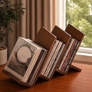 May include: Three wooden record holders, each angled, displaying vinyl records. The holders are made of dark wood and are positioned on a wooden surface. The records are visible, with album art and text. A plant and window are in the background.