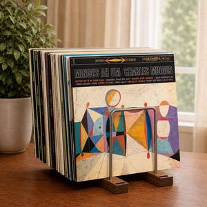 May include: A wooden and metal record holder displaying several vinyl records. The album cover visible features colorful geometric shapes and the text "MINGUS AH UM / CHARLES MINGUS". The holder is on a wooden surface.