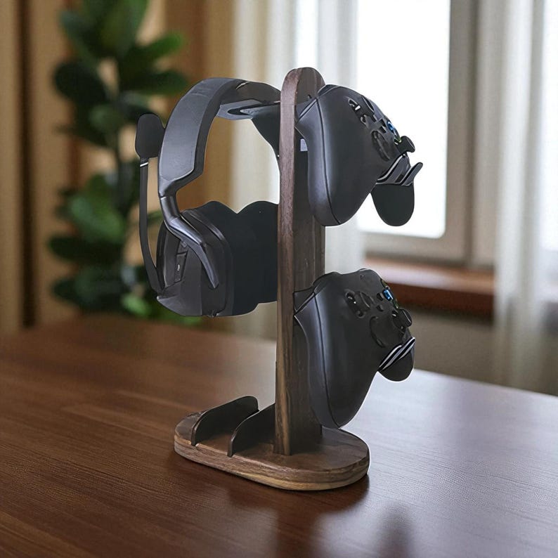 May include: A wooden gaming headset and controller stand. The stand is made of dark brown wood and holds a black gaming headset with a microphone and two black game controllers. The stand is on a brown surface.