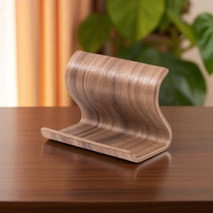 May include: A wooden phone stand with a curved, wave-like design. The stand is made of light brown wood with darker brown grain patterns. It has a small lip at the bottom to hold a phone and a curved back for support.