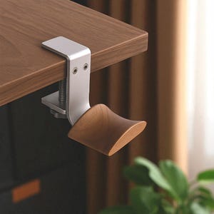 Peut inclure: Un support pour casque en métal argenté et bois, fixé à un bureau en bois. Le support comprend un berceau en bois incurvé et une pince métallique avec une vis. Il est conçu pour maintenir un casque.