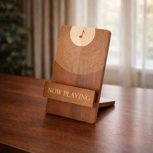 May include: Wooden phone stand with a musical note and vinyl record design. The stand is brown with the words "NOW PLAYING" engraved on the front. The stand is on a wooden surface.