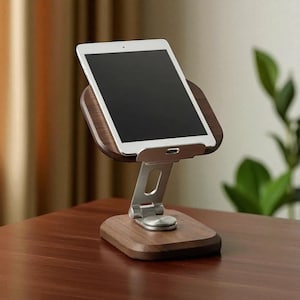 May include: A wooden tablet stand with a silver adjustable arm and base. The stand holds a white tablet with a black screen. The base and the tablet holder are made of dark brown wood.