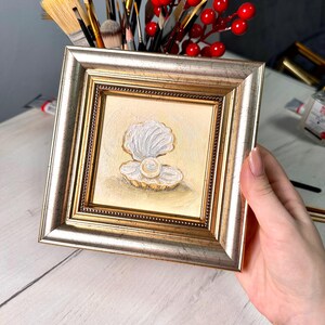 Könnte beinhalten: Gerahmtes Gemälde einer Perle in einer geöffneten Muschel. Das Kunstwerk hat einen goldenen Hintergrund und einen silber-goldenen Rahmen. Die Muschel ist weiß und die Perle hat eine cremeweiße Farbe. Das Gemälde wird von einer Person gehalten.