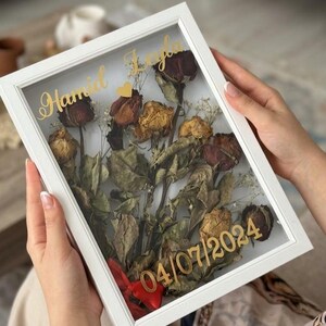 May include: A white framed shadow box with dried roses and leaves. Gold text reads "Hamid Leyla" and "04/07/2024". A red ribbon is tied at the bottom. The shadow box is held by a person.