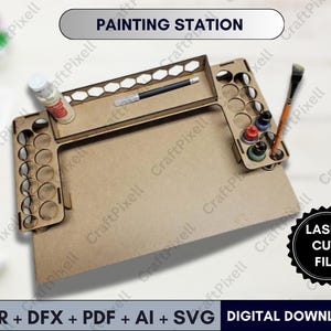 Może przedstawiać: Stacja malarska z drewna ciętego laserem z napisem "PAINTING STATION". Posiada wiele okrągłych i prostokątnych przegródek do przechowywania butelek z farbą, pędzli i innych materiałów artystycznych. Projekt obejmuje dużą płaską powierzchnię do malowania. Obraz zawiera również tekst "LASER CUT FILE" i "DIGITAL DOWNLOAD".