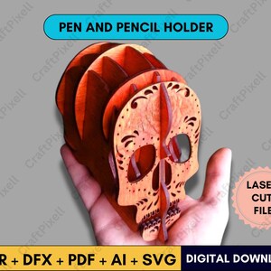 May include: A wooden pen and pencil holder in the shape of a skull, with intricate laser-cut details. The holder is a warm orange color and features the text "PEN AND PENCIL HOLDER". The design includes decorative patterns and is held in a hand.