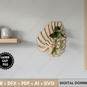 May include: A wooden monstera leaf-shaped wall planter with a glass vial holding a small plant. The planter is made of light-colored wood and is mounted on a gray wall. The image also includes a shelf with plants and the text "LASER CUT FILE".