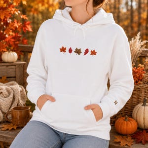 Könnte beinhalten: Ein weißer Kapuzenpullover mit einer horizontalen Reihe aus fünf gestickten Herbstblättern in Rot-, Orange- und Brauntönen auf der Brust. Der Hoodie verfügt über eine Kängurutasche vorne und einen kleinen Buchstaben N auf dem linken Ärmel.