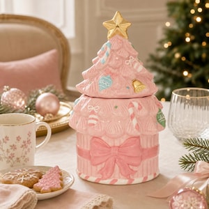 Op de afbeelding: Een keramische koekjespot in de vorm van een kerstboom met een pastelroze afwerking. Het deksel heeft een gouden ster als top en gegoten ornamenten. De basis is versierd met een grote roze strik en zuurstokstrepen langs de onderrand.